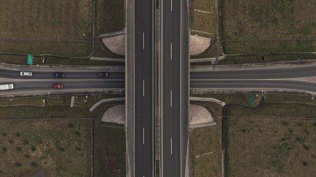 A top-down aerial view of a highway intersection with an overpass crossing above a main road. Several cars travel in both directions, surrounded by dry grassland and structured road layouts.