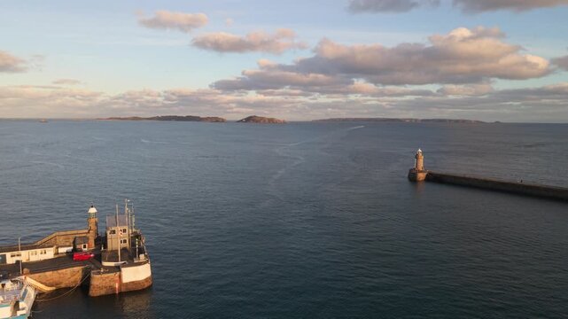 St Peter Port Harbour,Guernsey,reveal of harbour entrance passing berthed ferry in golden hour sun with Herm , Jethou and Sark beyond breakwater with calm sea and boat leaving port