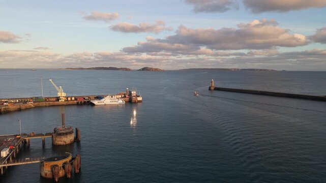 St Peter Port Harbour,Guernsey,reveal of harbour entrance passing berthed ferry in golden hour sun docks and ferry berths with Herm , Jethou and Sark beyond breakwater with calm sea and boat leaving
