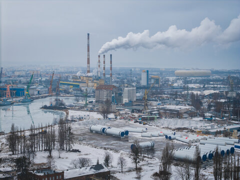Aerial view of industrial chimneys spewing plumes of smoke into the sky, contrasting against the snow-dusted ground and icy waters of the port, Gdansk, Poland.
