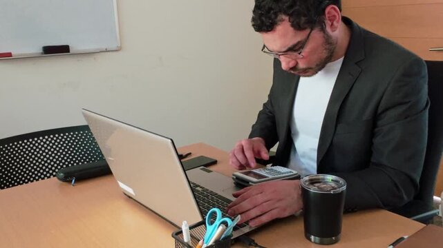 Diligent young man crunching numbers using a graphing calculator and laptop in an office setting.