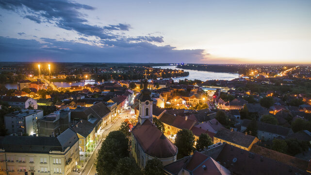 Aerial view of the illuminated bridge stretching across the wide river, the church standing tall amidst the red rooftops, Sremska Mitrovica, Vojvodina, Serbia.