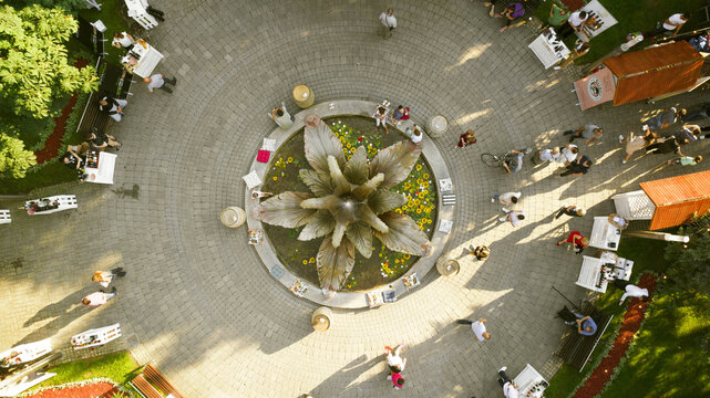 Aerial view of people gathering around a circular fountain with metallic leaves and yellow flowers, surrounded by tables and chairs, creating a vibrant social space. Sremska Mitrovica, Serbia.