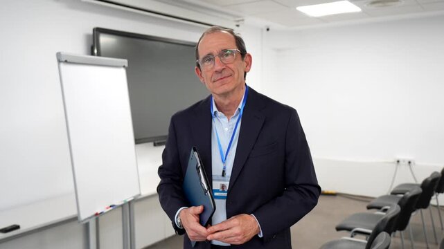 Confident senior medical professional with a clipboard standing in an empty hospital conference room, looking at the camera with a calm and reassuring expression before a medical seminar or meeting
