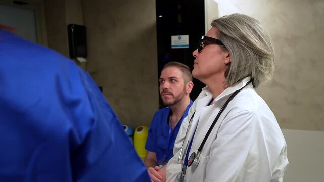 Mature female doctor with gray hair and glasses in lab coat and stethoscope speaking and gesturing while briefing younger male nurses in hospital staff room during clinical meeting