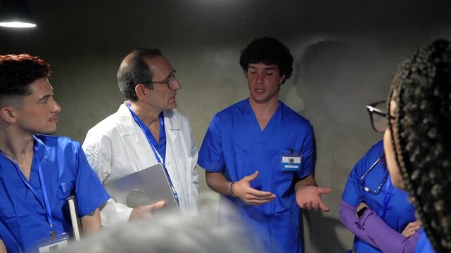Group of diverse medical students and a senior doctor gathered around a table for a briefing, with a young intern explaining a clinical case to his colleagues in a serious hospital setting