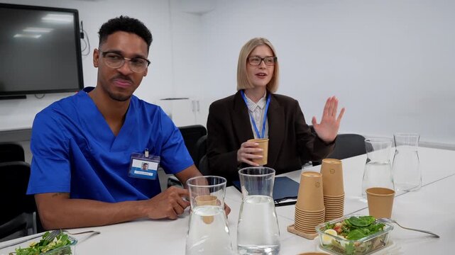 Diverse group of hospital staff nurses and doctors in scrubs sitting at a table during lunch break, eating salads, drinking water and enjoying a friendly conversation