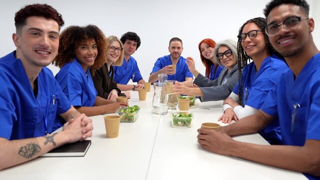 Diverse team of smiling doctors and nurses sitting around a table during a hospital meeting or lunch break, looking at camera in a bright conference room on white background
