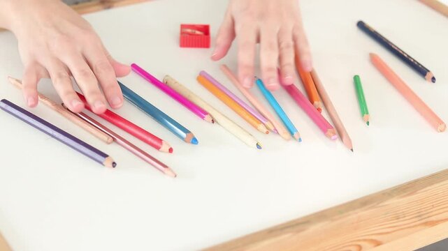 Hands touching scattered colored pencils with sharpener on white table