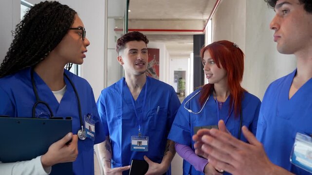 Group of four diverse young medical students in blue scrubs discussing a clinical case while standing together in a modern hospital corridor, collaborating on a diagnosis and sharing knowledge