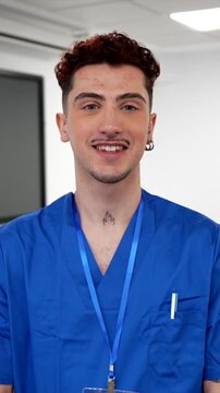 Confident young male medical professional with tattoos wearing a blue uniform and identification badge looking at the camera and smiling while standing in a modern clinic or hospital