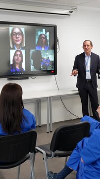 Male doctor leading a medical seminar. Teaching a class of students in scrubs while connecting with other participants remotely through a large video conference screen in a modern hospital classroom