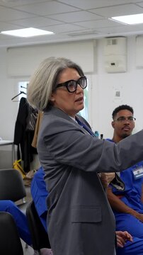 Senior female physician with grey hair and glasses speaking to a group of medical students during a professional training course, mentoring the next generation of healthcare professionals
