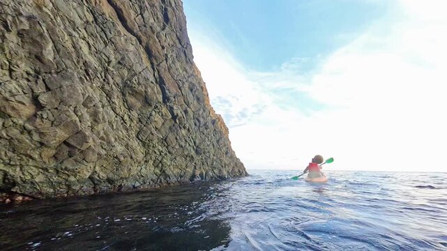 Kayaking cliff ocean adventurer in a red boat paddles along high rock walls on a calm sunny day for summer exploration