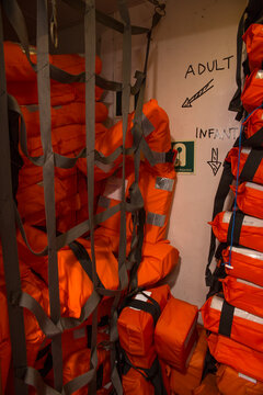 Tall stack of bright orange life jackets stored in a compartment on a ship. The life vests are packed tightly with visible straps and reflective tape, ready for emergency use during a maritime voyage.