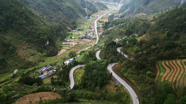 Aerial drone footage captures a winding mountain road with sharp hairpin turns through a green valley with a small mountain village surrounded by steep hills in northern Vietnam