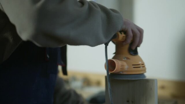 A person uses an orbital sander to smooth the surface of a wooden block. The craftsman diligently works on refining the timber in a workshop setting.