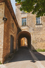 Fototapeta premium A picturesque stone archway frames a serene view of a narrow cobblestone street, illuminated by bright sunshine in a quaint French village. Old buildings whisper stories of the past