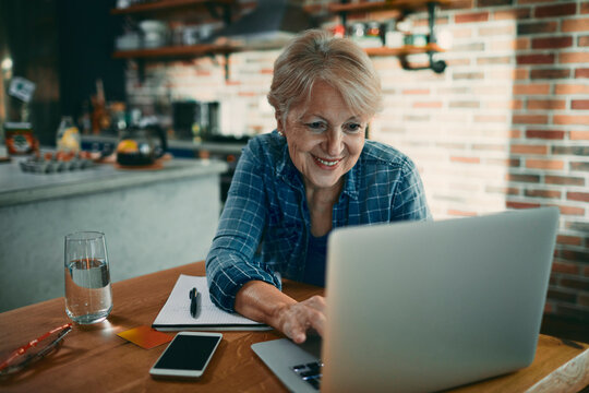Smiling senior woman working on laptop at home kitchen