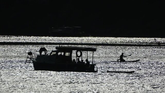 Boat paddleboard water. Silhouetted person paddling a stand up paddleboard across shimmering water near tour boats in high contrast lighting