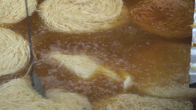 Close-up of traditional kataifi pastry nests being deep-fried in hot, bubbling oil, with a cook using a utensil to turn them until they achieve a perfect golden-brown color