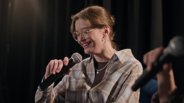 Panning shot of smiling multiethnic young adult woman sharing funny stories on stage while participating in comedy panel show with female comedians under spotlight