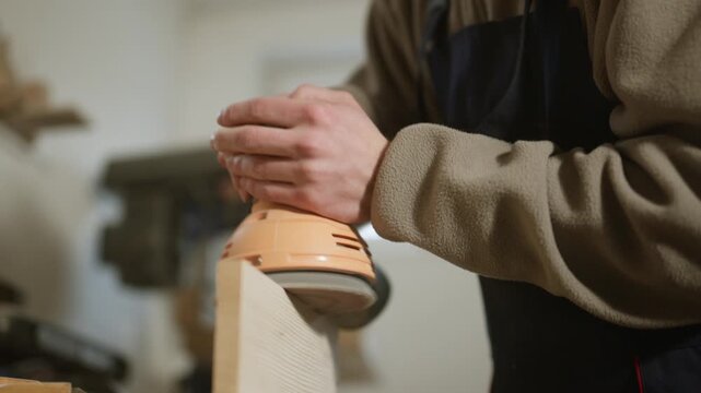 A carpenter meticulously sands a wooden plank with an orbital sander in a bright workshop. This close-up shot shows the focused work of preparing wood for a project.