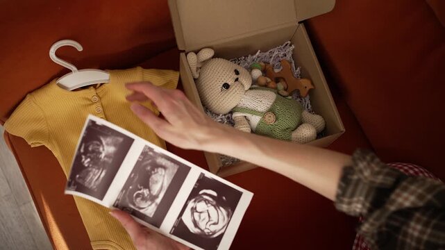 Pregnant woman preparing for childbirth, looking at an ultrasound image of her unborn baby, tenderly holding a yellow newborn bodysuit, with a gift box of toys nearby