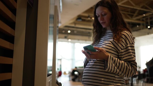 Expectant mother standing at a self-service terminal, using the interactive touchscreen and her smartphone to check in for a flight, buy tickets, or make a contactless payment