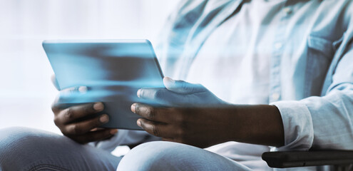 Fototapeta premium An adult African American man in a wheelchair uses a tablet to chat and work remotely. He sits in a living room during the COVID pandemic, engaging in online activities.