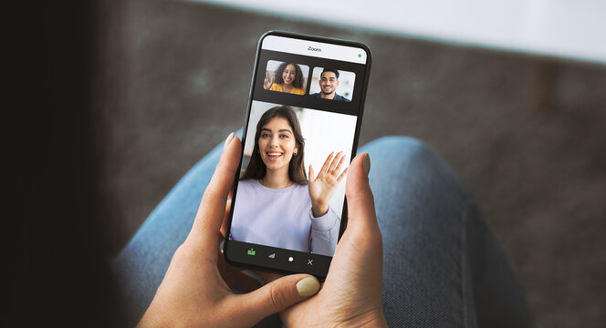 A millennial lady is holding a smartphone in a living room. She is engaging in a remote work call with two other people on the screen. The setting shows a relaxed home environment.