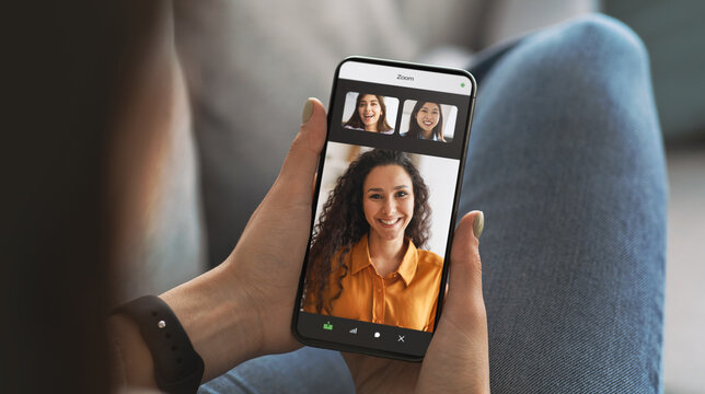 Young lady sits on a sofa while holding a smartphone displaying a video call. She engages with a friend on a virtual platform during a time of self-isolation at home.