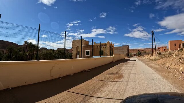 Driving through moroccan village buildings and oasis in the ziz valley on a scenic desert road trip under a blue sky pov