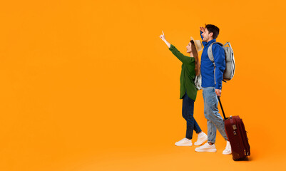 Fototapeta premium A couple stands on an orange background. They seem excited and point at something ahead. They carry a suitcase and have backpacks. Their expressions show anticipation for their journey.