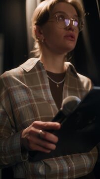 Vertical handheld shot of focused young adult female comic with glasses revising speech on clipboard feeling nervous while standing backstage