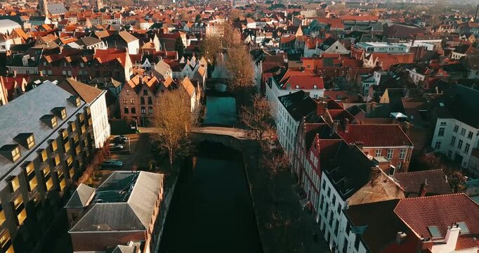 Breathtaking aerial drone footage of the historic canals in Bruges, Belgium. Charming old buildings with red-tiled roofs line the waterway, crossed by a picturesque stone bridge under warm sunlight.