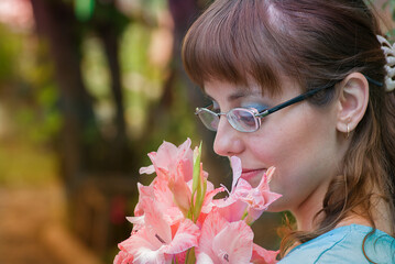 Close-up profile portrait of a woman with glasses gently smelling a bouquet of pink gladiolus flowers.Concept of sensory pleasure, spring bloom, and mindful living. © Yelena