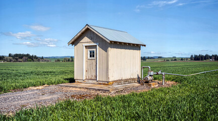 Obraz premium Irrigation well pump and small pump house in a green agricultural field. Farm water supply system under a blue sky. Rural landscape with farming infrastructure