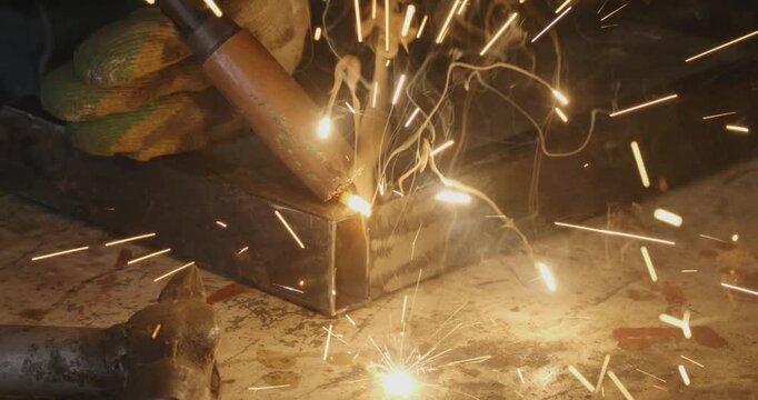 Professional welder joining metal profiles with electric arc welding, creating bright sparks and smoke in a workshop. The process shows the precise creation of a metal frame corner joint.