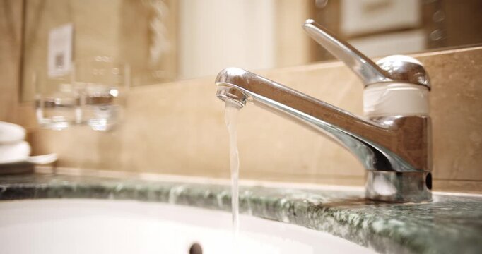 A steady, weak stream of water flows from a sleek silver faucet into a green marble vanity sink. Close up shot, shallow depth of field, real time video