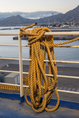 Fototapeta premium A thick, yellow mooring rope coiled around a white metal railing on a ship's deck. In the background, a harbor and misty mountains are visible under a soft sky, capturing a quiet maritime moment.