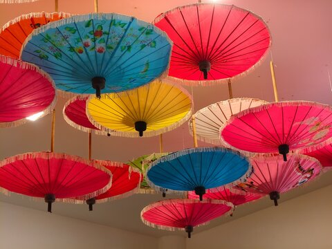 A vibrant ceiling display of traditional handcrafted Asian oil-paper umbrellas in various colors like pink, blue, and yellow, featuring delicate floral patterns and fringe details.