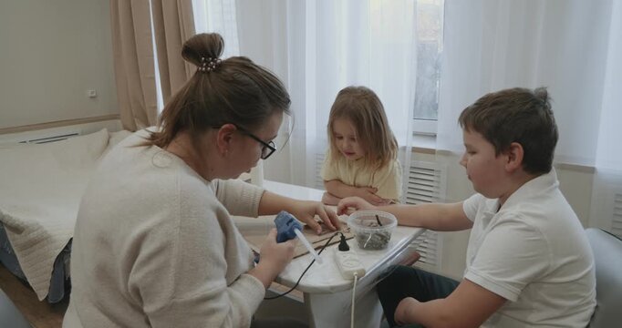 Creative family time at home with mother, son and daughter working together on a diy project, using a hot glue gun and wooden sticks to build something new and imaginative at the table.