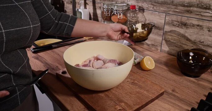Female hands adding a creamy mayonnaise and spice marinade from a small glass bowl to raw chicken drumsticks in a large bowl, preparing a delicious homemade meal in a modern kitchen.