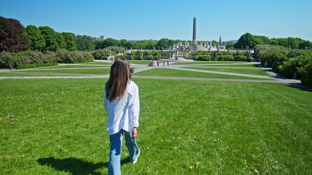 Tourist woman walking through green lawn toward the Monolith sculpture in Frogner Park. Monolith column and sculpture park landscape during summer in Oslo, Norway.