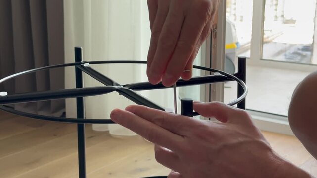 Hands tighten bolt using hex key on metal frame during table assembly. Fingers screw fastener into structure while building furniture at home. Concept diy, setup, interior..