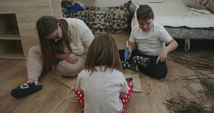 Young family, including a mother, son, and daughter, sitting on the floor at home engaged in a creative diy project, crafting frames from natural materials like small twigs and cardboard.