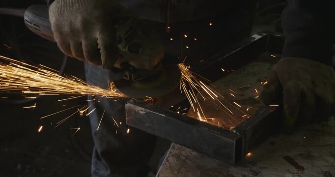 Professional craftsman in protective gloves using an angle grinder to clean and smooth a metal profile pipe, creating bright sparks in a workshop before the welding process.