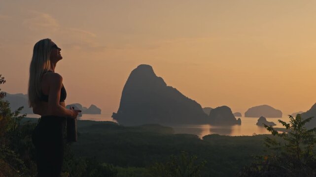 Fit woman in activewear walks into frame, drinks from a water bottle, and admires limestone sea cliffs under a golden sky. Peaceful fitness lifestyle in a scenic coastal setting