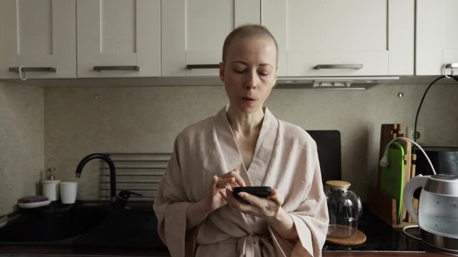 shaved head woman holds a smartphone and gazes to the side in a home kitchen, reflective pause about mental health, coping with illness, resilience, and everyday recovery life